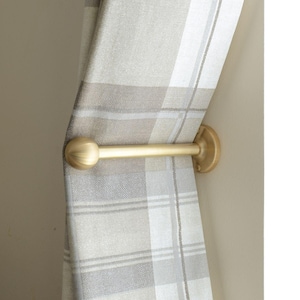 Puede incluir: Un recogedor de cortina dorado con un remate redondo y una placa de pared. Sujeta una cortina a cuadros beige y gris. La cortina está hecha de una tela tejida.