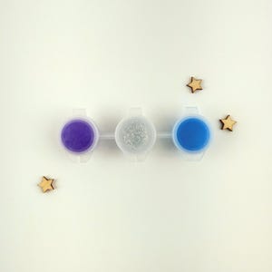 May include: Three small paint pots with lids, arranged in a row. The pots contain purple, silver glitter, and blue paint.