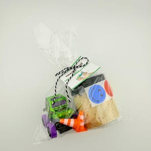 Puede incluir: Una bolsa de celof&aacute;n transparente atada con hilo blanco y negro, que contiene un cami&oacute;n de juguete verde y morado, conos de tr&aacute;fico naranjas y un frasco con pegatinas de caritas sonrientes. Una etiqueta dice "&iexcl;Gracias por decir buenos momentos!"