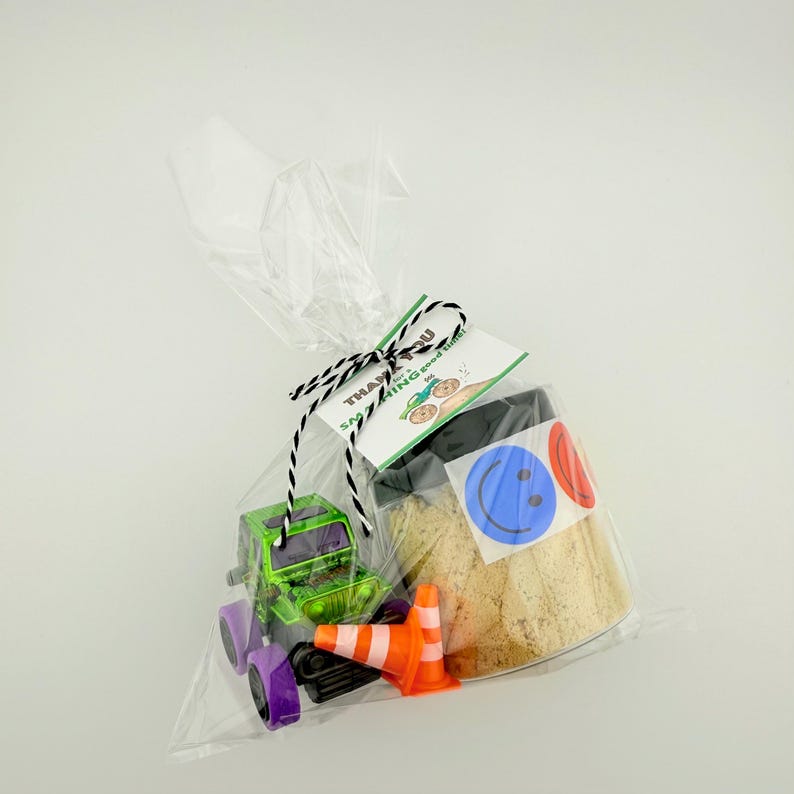 Puede incluir: Una bolsa de celof&aacute;n transparente atada con hilo blanco y negro, que contiene un cami&oacute;n de juguete verde y morado, conos de tr&aacute;fico naranjas y un frasco con pegatinas de caritas sonrientes. Una etiqueta dice "&iexcl;Gracias por decir buenos momentos!"