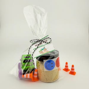 Puede incluir: Una bolsa de regalo transparente con un cami&oacute;n monstruo verde de juguete, conos de tr&aacute;fico naranjas y un tarro con tapa negra. El tarro tiene una pegatina de carita sonriente azul y una pegatina roja. La etiqueta dice "Thank you for a SMASHING good time!"