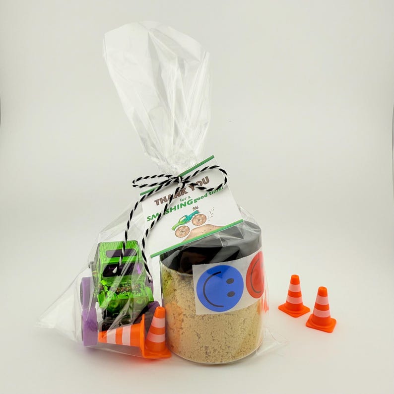 Puede incluir: Una bolsa de regalo transparente con un cami&oacute;n monstruo verde de juguete, conos de tr&aacute;fico naranjas y un tarro con tapa negra. El tarro tiene una pegatina de carita sonriente azul y una pegatina roja. La etiqueta dice "Thank you for a SMASHING good time!"