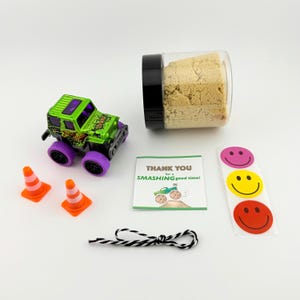 Puede incluir: Un juguete de cami&oacute;n monstruo verde y negro con ruedas moradas, dos conos de tr&aacute;fico naranjas, un frasco con contenido beige, una hoja de pegatinas con caras sonrientes, una tarjeta que dice "THANK YOU for a SMASHING good time!" y una cuerda en blanco y negro.