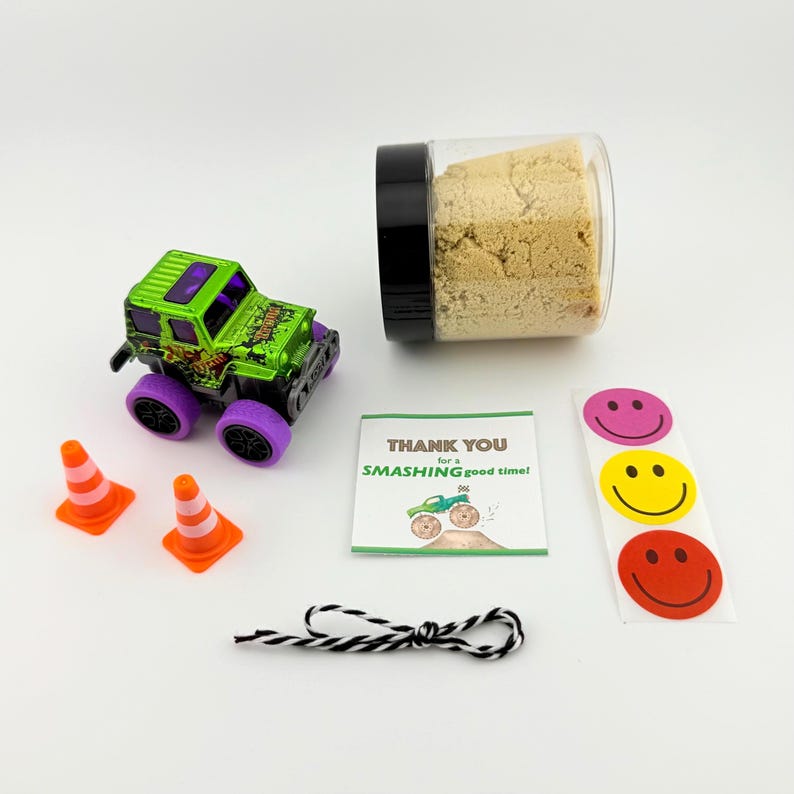 Puede incluir: Un juguete de cami&oacute;n monstruo verde y negro con ruedas moradas, dos conos de tr&aacute;fico naranjas, un frasco con contenido beige, una hoja de pegatinas con caras sonrientes, una tarjeta que dice "THANK YOU for a SMASHING good time!" y una cuerda en blanco y negro.