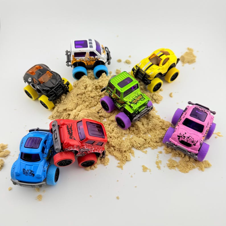 Puede incluir: Una colecci&oacute;n de camiones monstruo de juguete coloridos en varios colores, incluyendo azul, rojo, rosa, verde, amarillo, negro y blanco. Los camiones est&aacute;n colocados sobre un lecho de arena, mostrando sus grandes ruedas y dise&ntilde;os robustos.