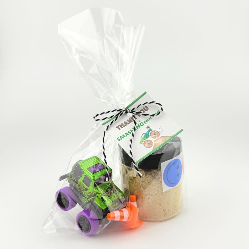 Puede incluir: Una bolsa de celof&aacute;n transparente atada con hilo blanco y negro, que contiene un juguete de cami&oacute;n monstruo verde y negro con ruedas moradas, un cono de tr&aacute;fico naranja y un frasco con una pegatina de carita sonriente azul. La etiqueta dice "Thank you for a smashing good time!"