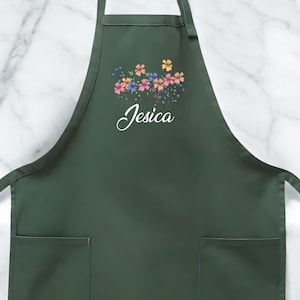 May include: Dark green apron with two front pockets and a floral design. The apron has a cluster of colorful flowers at the top and the name "Jesica" in cursive script.