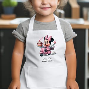May include: A white apron featuring a graphic of Minnie Mouse riding a pink scooter while holding a cupcake and a fork. Below the graphic, the text reads Linda and cookie baker in black script and sans-serif font. The apron has a front pocket.