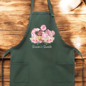 May include: Dark green apron with two front pockets. The apron features a watercolor design of sweets, including a donut, cupcake, and heart-shaped candies, surrounded by pink roses. The text "Susan's Sweets" is printed below the design.