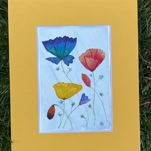 Può includere: Un dipinto ad acquerello di papaveri colorati nei toni del blu, rosso e giallo. L'opera è incorniciata in bianco e in una cornice giallo brillante. Il dipinto è su uno sfondo azzurro.