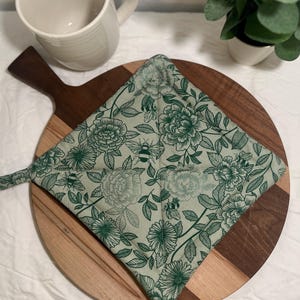 May include: A square, quilted pot holder with a green floral and bee pattern. The pot holder has a loop for hanging and is placed on a wooden cutting board. The design features large flowers and leaves with a light green background.