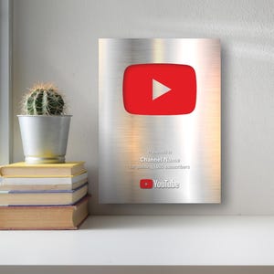 Puede incluir: Un premio de botón de reproducción de YouTube plateado con un logotipo de botón de reproducción rojo. El premio se otorga a un canal por superar los 1.000 suscriptores. Un pequeño cactus en una maceta de metal y una pila de libros están al lado del premio.