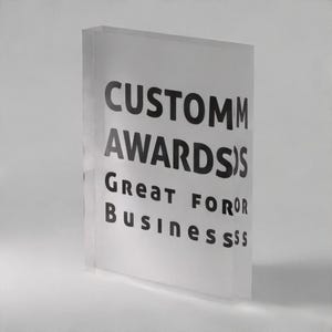 May include: A rectangular clear acrylic block award featuring black printed text that reads CUSTOM AWARDS GREAT FOR BUSINESS. The block has a thick, transparent profile with a smooth, polished finish and a flat base for upright display on surfaces.