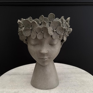 May include: A grey concrete planter shaped like a human head with closed eyes and a flower crown. The planter is on a white surface.