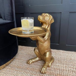 May include: A gold-coloured dog-shaped side table featuring a seated canine figure holding a round, flat tray. A clear glass filled with a carbonated beverage, ice cubes, and a yellow lemon slice sits on the tray surface of the metallic sculpture.