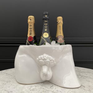 May include: White ceramic champagne bucket shaped like a human torso. Three bottles of champagne are inside the bucket, two are Moët & Chandon and one is Taittinger Brut.