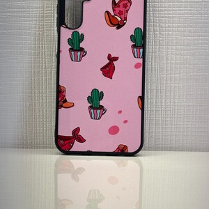 May include: A pink phone case with a black border. The case has a pattern of cacti, cowboy boots, and bandanas.