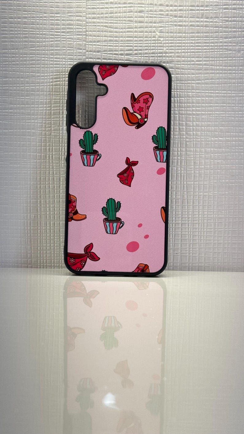 May include: A pink phone case with a black border. The case has a pattern of cacti, cowboy boots, and bandanas.