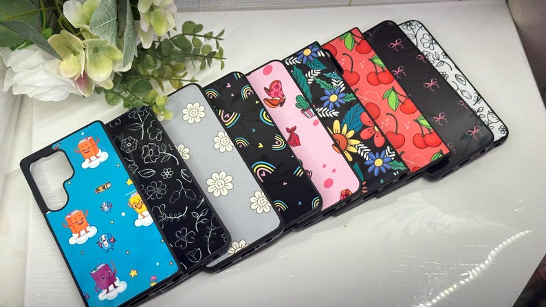 May include: A set of eight phone cases with different colorful and whimsical designs. The cases are made of a hard plastic material and have a glossy finish. The designs include rainbows, flowers, cherries, and other cute patterns.