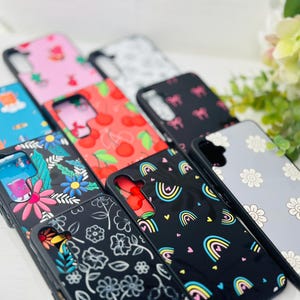 Puede incluir: Una colección de fundas para teléfono con varios diseños coloridos y estampados. Las fundas presentan diferentes patrones, incluyendo cerezas, flores, arcoíris y diseños abstractos. Todas las fundas son negras con una capa protectora transparente sobre el diseño.