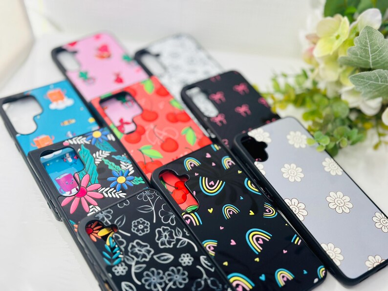 May include: A collection of phone cases with various colorful and patterned designs. The cases feature different patterns, including cherries, flowers, rainbows, and abstract designs. The cases are all black with a clear protective layer over the design.