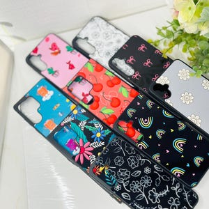 May include: A collection of colorful phone cases with various patterns, including cherries, rainbows, flowers, and abstract designs. The cases are all black with a clear back.