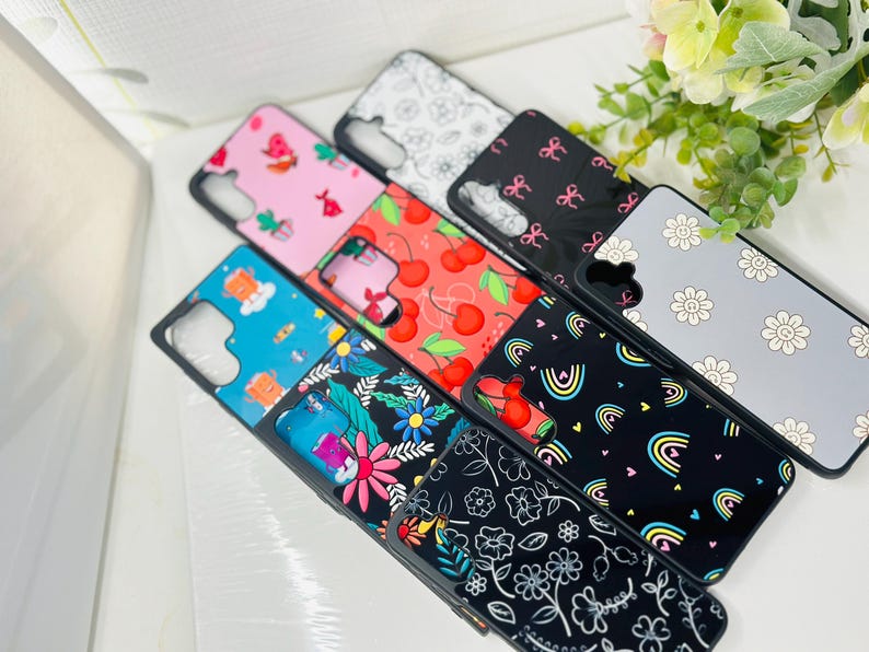 May include: A collection of colorful phone cases with various patterns, including cherries, rainbows, flowers, and abstract designs. The cases are all black with a clear back.