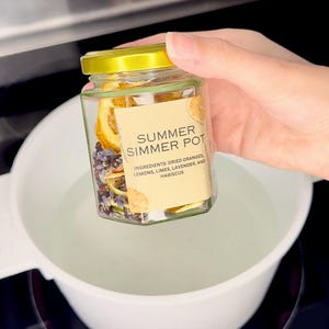 May include: A glass jar filled with dried citrus fruits, lavender, and hibiscus. The jar has a yellow lid and a label that reads "Summer Simmer Pot" with the ingredients listed below.
