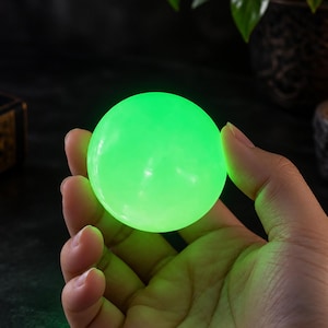 Può includere: Un oggetto sferico liscio che emette una luce verde brillante e vibrante. La superficie della sfera è uniforme e appare solida, proiettando una luce soffusa sull'area circostante. L'oggetto è tenuto nel palmo di una mano.