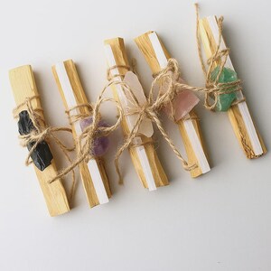 Puede incluir: Seis paquetes de palos de Palo Santo, cada uno adornado con un cristal diferente. Los cristales incluyen piedras negras, moradas, transparentes, rosas y verdes, todos atados con hilo. Los palos son de color marrón claro con una franja blanca.