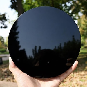 May include: A round, black, polished obsidian stone held in a hand. The stone reflects the surrounding trees and sky, creating a mirror-like effect. The obsidian has a smooth, glossy surface.