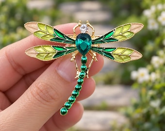May include: A dragonfly brooch featuring a gold-tone metal frame, four wings with green and light yellow enamel inlay, a teardrop-shaped dark green crystal body, a small clear round crystal head, and a tail made of small green beads.