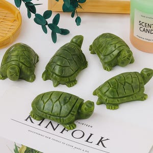 Op de afbeelding: Vijf groene schildpadbeeldjes met gedetailleerde schaalpatronen zijn gerangschikt op een wit oppervlak. Het woord "KINFOLK" is zichtbaar op een tijdschrift. Een kaars en decoratieve bladeren zijn op de achtergrond.