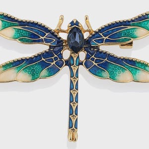 Op de afbeelding: Een goudkleurige libellebroche met blauwe, turquoise en crèmekleurige geëmailleerde vleugels. De libelle heeft een donkerblauwe edelsteen in het midden en een sluiting met speld aan de achterkant. De vleugels hebben een gedetailleerd, getextureerd ontwerp.