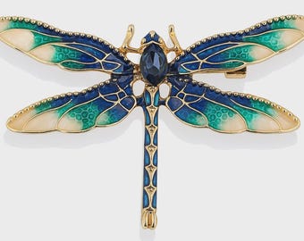May include: A gold dragonfly brooch with blue, teal, and cream enamel wings. The dragonfly has a dark blue gemstone at its centre and a pin closure on the back. The wings have a detailed, textured design.
