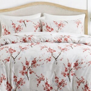 Pode incluir: Conjunto de roupa de cama branco com um padrão floral de flores vermelhas e galhos marrons. O conjunto inclui uma capa de edredom e duas fronhas, todos com o mesmo design. A cama é feita com uma cabeceira de madeira clara.