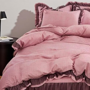 May include: A bed dressed with a pink duvet cover and matching pillowcases. The bedding features a textured design and is trimmed with a darker brown frill. A black lamp and side table are in the background.