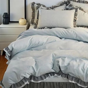 Puede incluir: Una cama con un edredón azul claro y fundas de almohada a juego. La ropa de cama tiene un ribete gris con volantes. Una mesita de noche blanca con tres cajones y un jarrón negro son visibles en el fondo. La habitación tiene una estética moderna y minimalista.
