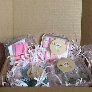 May include: A cardboard box filled with pink shredded paper and several clear plastic containers. The containers hold artificial nails with various designs, including floral patterns and landscape scenes. Each container has a small, round label with handwritten text.