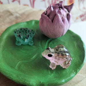 Puede incluir: Una hoja de nenúfar de cerámica verde sostiene dos pequeñas figuras de rana y una flor de loto púrpura. Una rana es verde con manchas negras, la otra es marrón y bronceada. El loto es de color púrpura apagado.