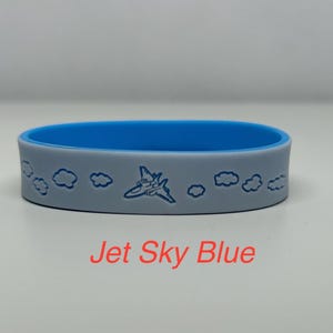 May include: A blue and gray silicone bracelet with a jet plane and clouds design. The text "Jet Sky Blue" is printed below the bracelet.