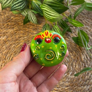 Può includere: Un oggetto decorativo rotondo di colore verde brillante che presenta un volto stilizzato con grandi occhi neri, un piccolo naso rosso e dettagli dipinti in giallo, tra cui una cresta, spirali e piccoli motivi a stella sulla superficie.