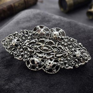 Könnte beinhalten: Eine ovale Haarspange aus silberfarbenem Metall mit einem komplexen Filigran-Design, durchbrochenen Mustern, texturierten Oberflächen und einem zentralen länglichen Motiv. Das Accessoire hat ein poliertes Finish mit dunklen Vertiefungen.