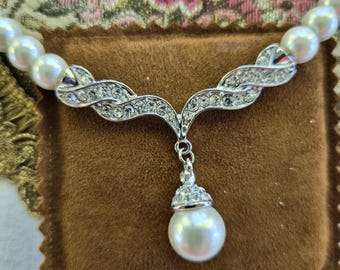 May include: A necklace featuring a strand of white pearls connected to a silver-toned metal centerpiece. The centerpiece is curved, encrusted with clear rhinestones, and supports a single dangling white pearl pendant topped with a rhinestone cap.