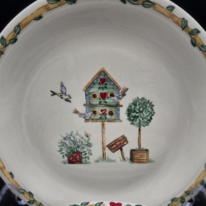 May include: Two white ceramic plates with a birdhouse design. The plates feature a border of vines, leaves, and red hearts. The center of the plates depicts a birdhouse, birds, and a small tree. The plates are on a clear stand.