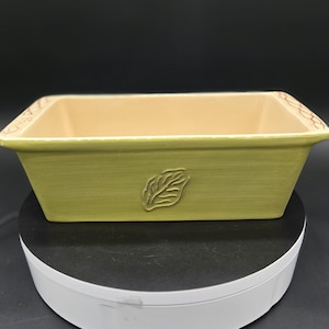 May include: A rectangular ceramic baking dish with a light green exterior and a beige interior. A leaf design is embossed on the front. The rim has a decorative pattern. The dish is on a white rotating platform.