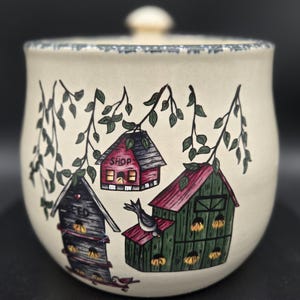 May include: Cream-colored ceramic jar with a lid, featuring a hand-painted design of birdhouses and foliage. The birdhouses are in shades of red, green, and black. The jar has a blue patterned rim and a small knob on the lid.