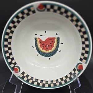 May include: White ceramic bowl with a watermelon design. The bowl features a black and white checkered border, a green trim, and a watermelon slice in the center. Small watermelon slices are also on the rim. The bowl is displayed on a clear stand.