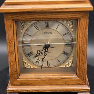 May include: A wooden quartz clock with a rectangular frame and a gold-colored handle. The clock face has Roman numerals and the brand name "LINDEN". The clock is made of light brown wood.