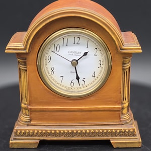 May include: A wooden Danbury clock with a white clock face and gold accents. The clock has Roman numerals and a quartz movement. The clock is a decorative item for a desk or shelf.